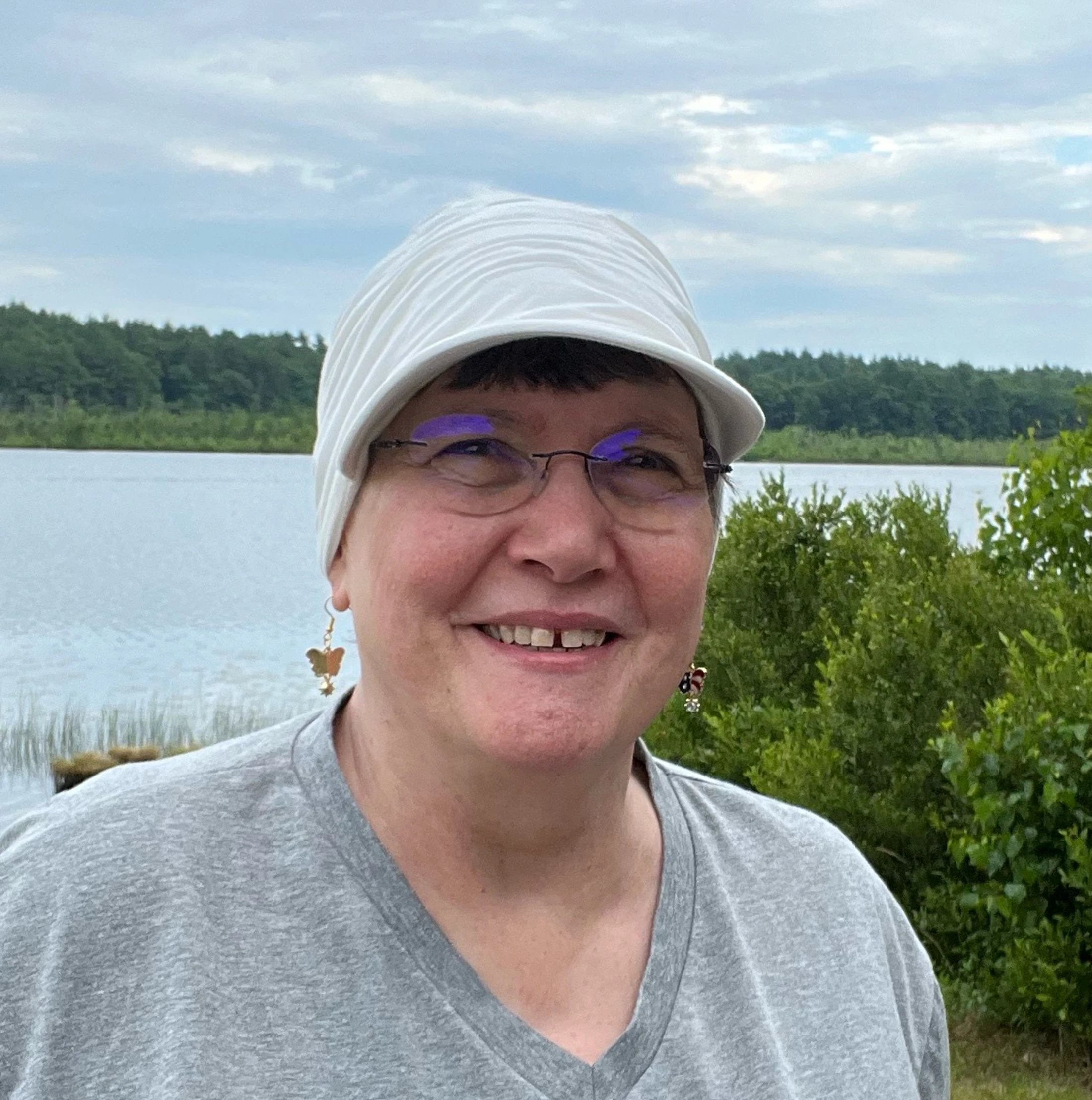 A headshot of a smiling woman wearing a white striped hat, glasses, and a grey t-shirt. She is standing outdoors with a lake and trees in the background.