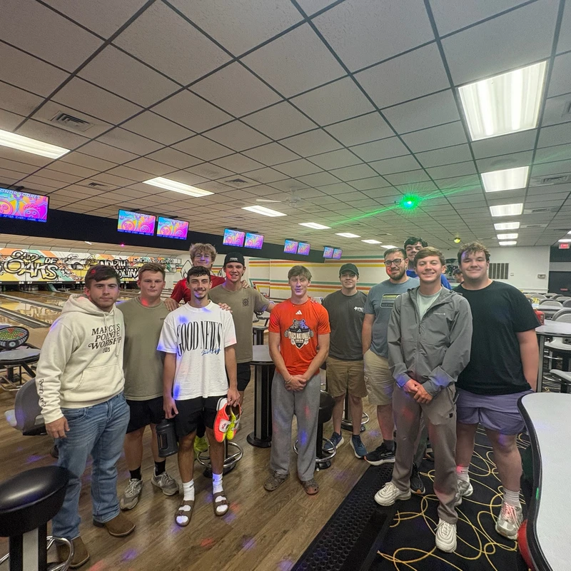 A group of eleven young men pose for a photo in a bowling alley, standing on the wood-look floor in front of the lanes and seating area.
