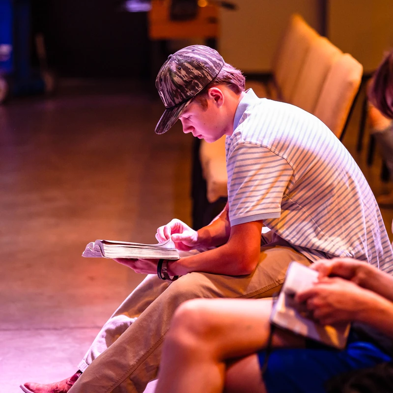 A young man in a striped shirt and camo hat sits in a row of chairs, looking down and reading from a book in his lap under warm, purple-toned lighting.
