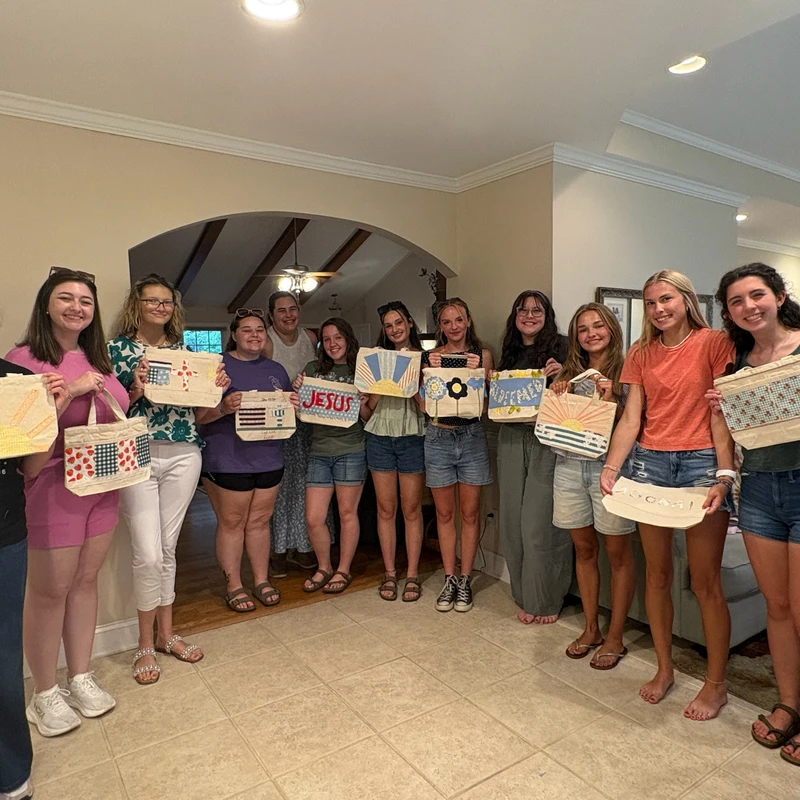 A group of eleven young women stand together in a home, smiling and proudly displaying the decorative fabric tote bags they have crafted.