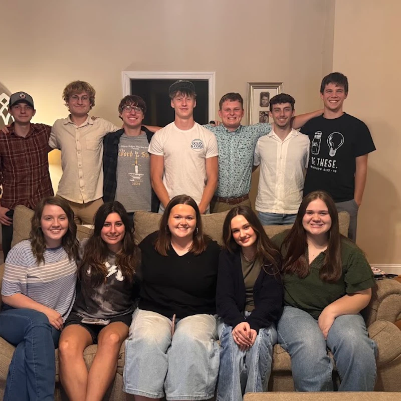 A group of twelve young adults smile for a photo. Five young women are seated on a couch in front of seven young men who are standing behind them.
