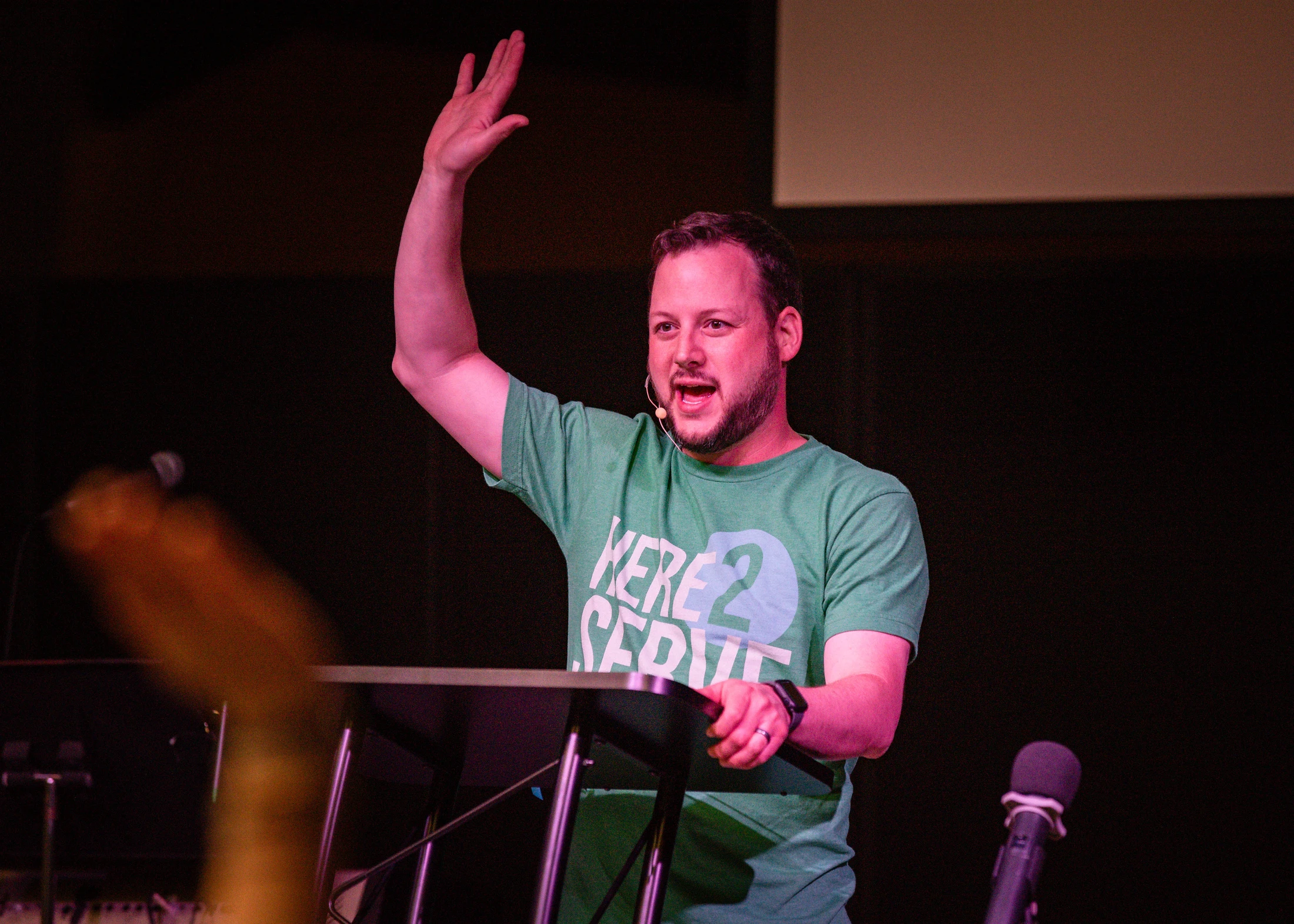 A man in a green t-shirt that reads 'Here 2 Serve' stands at a lectern on a stage, raising his hand as he speaks into a small microphone.