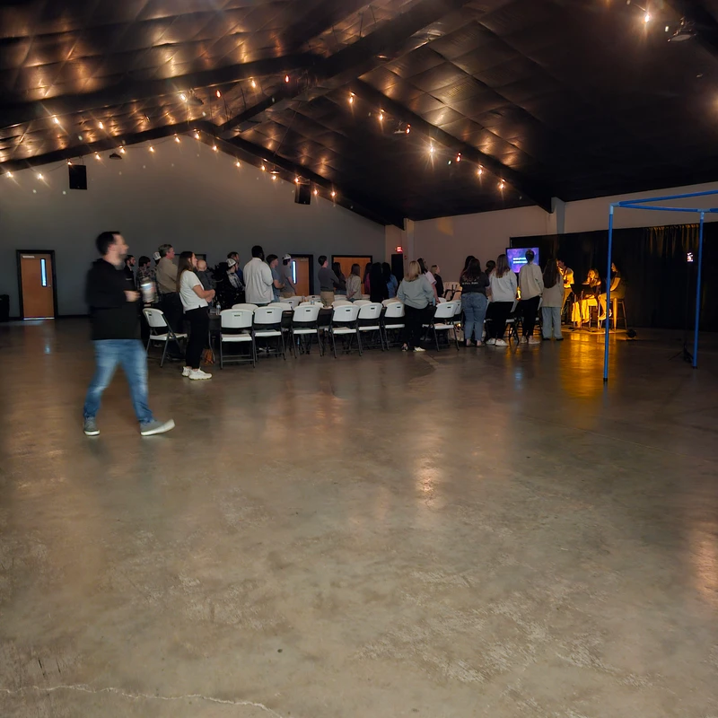 A wide shot of a large, dimly lit hall with a high ceiling and string lights. A group of people are gathered in the background for an event or meeting.