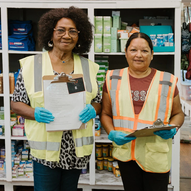Two women in yellow safety vests and blue gloves stand in front of stocked shelves in a warehouse, holding clipboards and smiling at the camera.