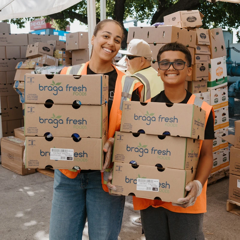 A smiling woman and a young boy in orange vests hold stacks of cardboard food boxes, with many more boxes stacked behind them at an outdoor charity event.