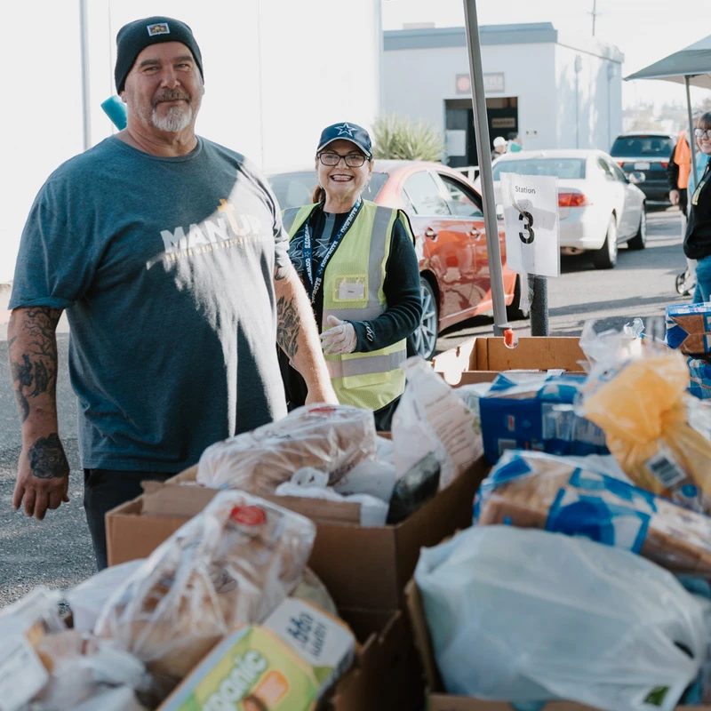 A smiling man and woman volunteer at a food drive, standing behind tables laden with boxes of donated food, with cars lined up in the background.