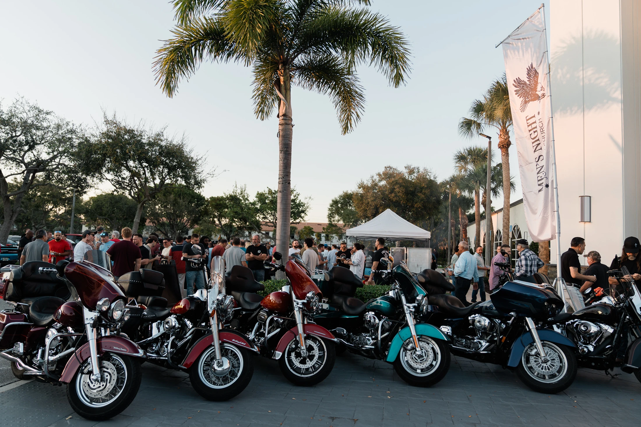 A row of colorful motorcycles is parked in the foreground. In the background, a crowd of men socializes at an outdoor 'Men's Night' event.
