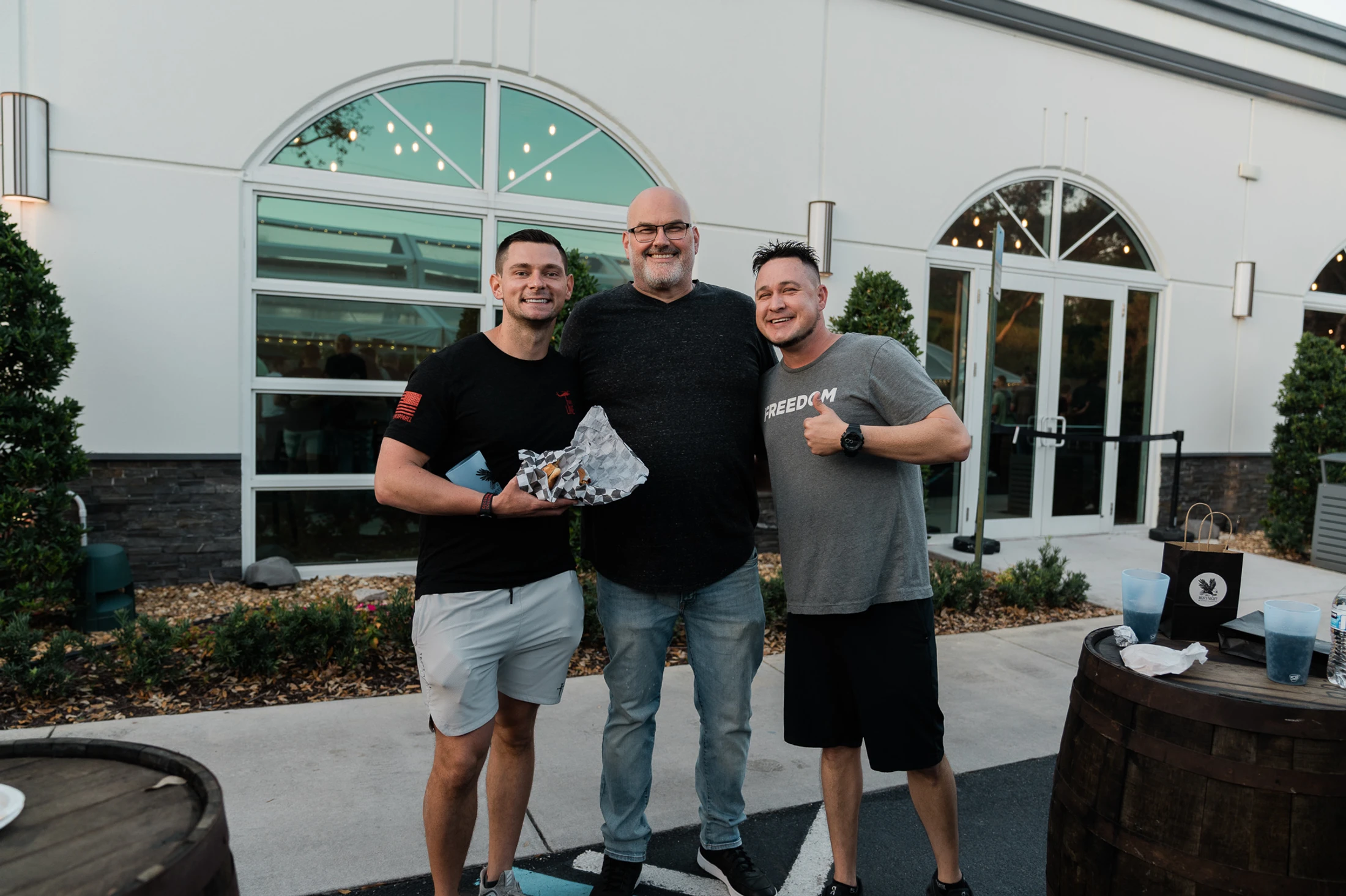 Three men smile for a photo outdoors in front of a white building. The man on the right gives a thumbs-up, and the man on the left holds a sandwich.