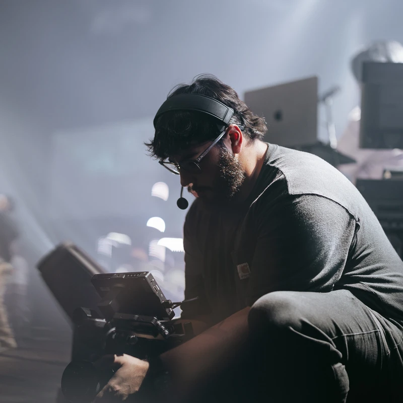 A bearded cameraman wearing headphones and glasses kneels to operate a video camera on a hazy set with bright spotlights in the background.