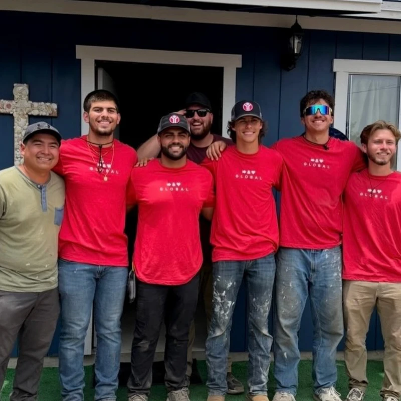 A group of seven smiling men stand arm-in-arm for a photo in front of a blue building. Most wear matching red t-shirts and have paint on their pants.