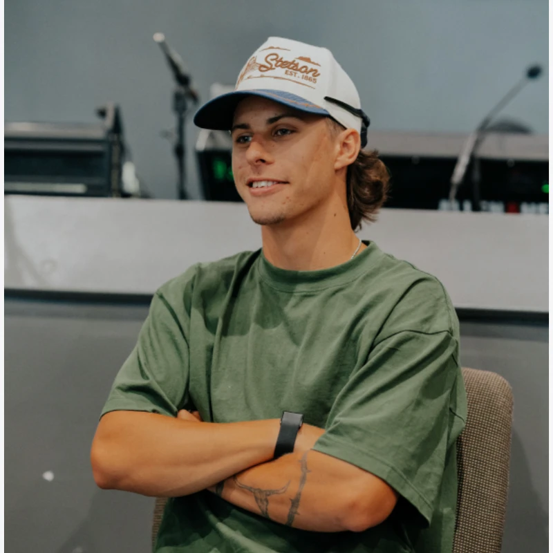 A young man with a mullet hairstyle sits in a chair, wearing a green t-shirt, jeans, and a Stetson hat. He has a tattoo on his arm and sits with them crossed.