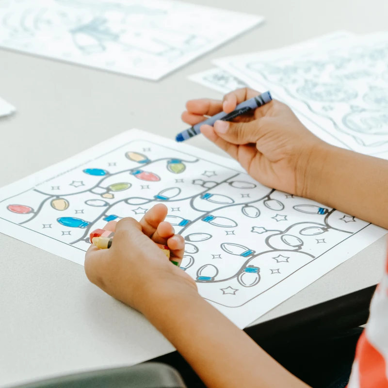 A close-up of a child's hands holding a blue crayon over a coloring page with a pattern of Christmas lights and stars on a white table.