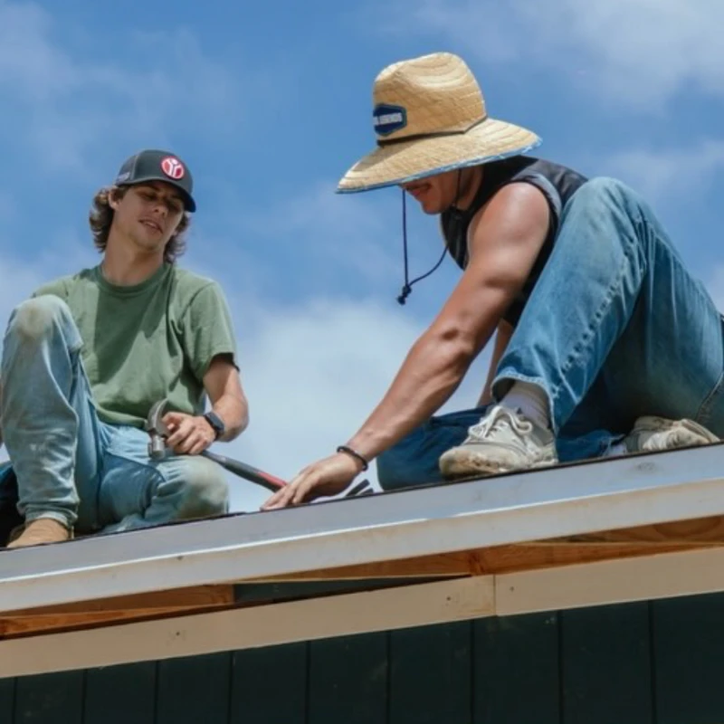 A low-angle shot of two young male workers sitting on the peak of a roof on a sunny day. One worker holds a hammer while the other points to the roof.