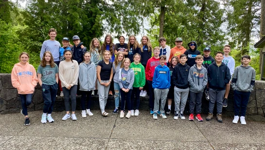 A large group of young students, boys and girls, stand in two rows on and in front of a stone wall, posing for a photo with green trees behind them.