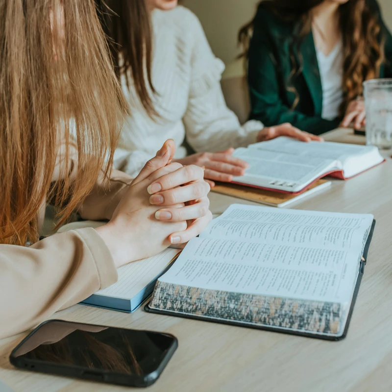 Three women sit at a light wood table with open books, possibly Bibles, in front of them. The focus is on the woman in the foreground with her hands clasped.