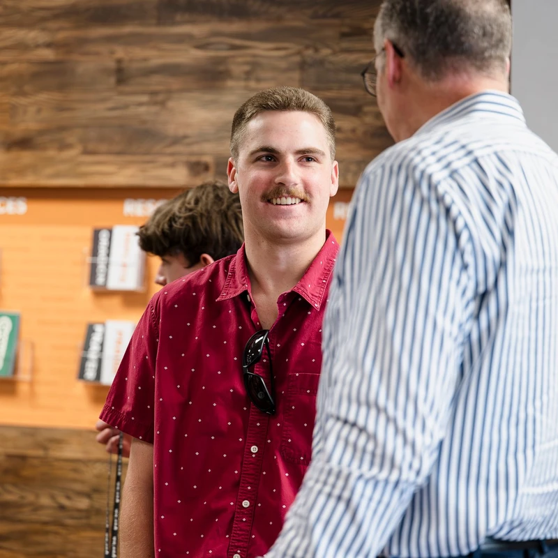 Un joven con bigote, que lleva una camisa roja estampada, sonríe mientras habla con otro hombre que lleva una camisa a rayas. Están de pie en el interior de un vestíbulo.