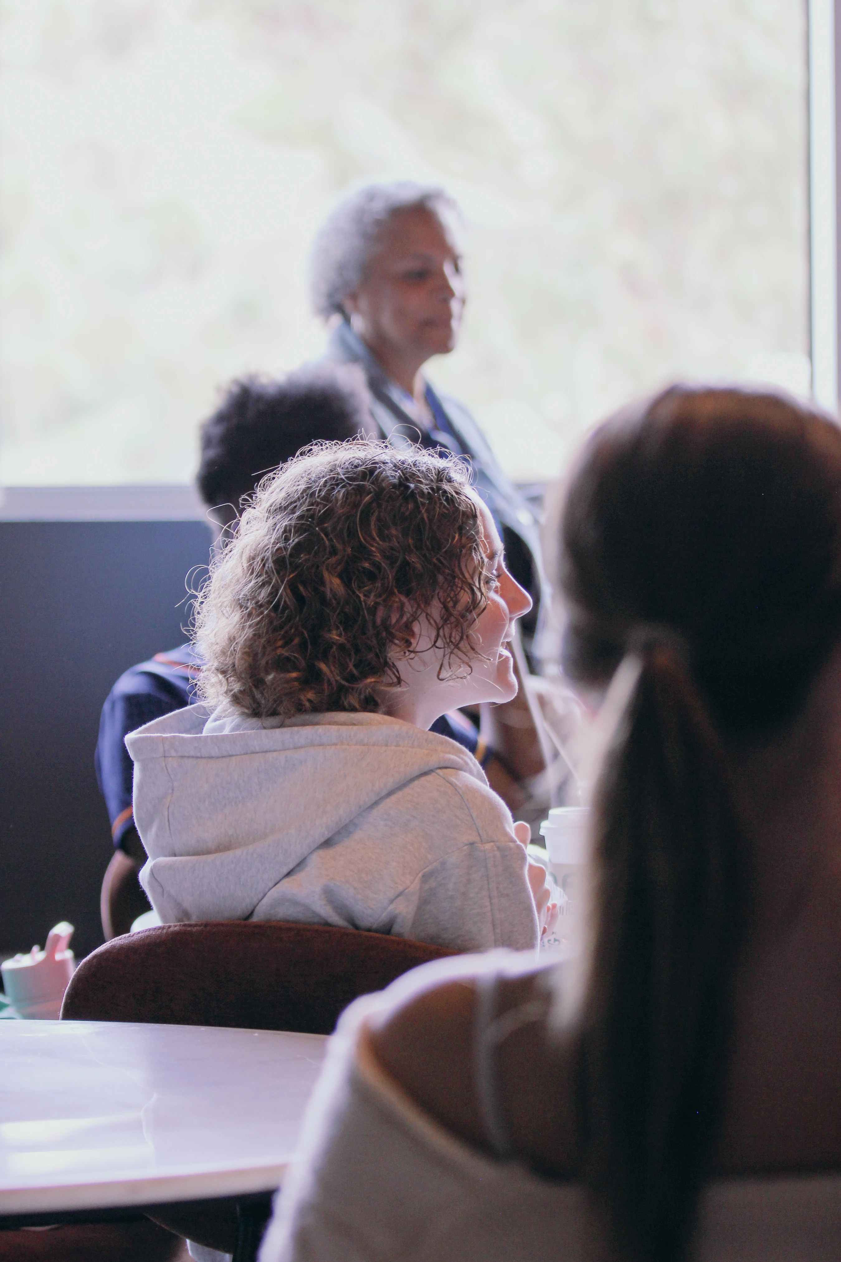 A young woman with curly brown hair, wearing a grey hoodie, smiles while sitting in a group. An older woman is visible in the background, out of focus.