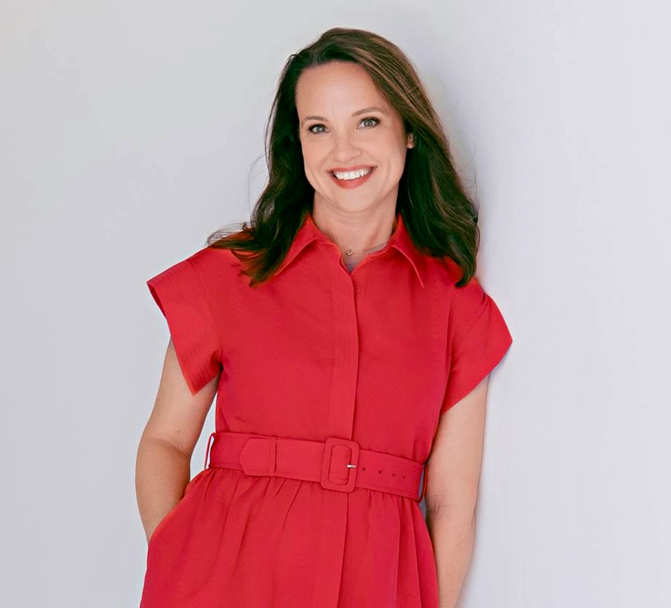 A professional headshot of a smiling woman with brown hair wearing a short-sleeved, collared red dress with a belt, leaning against a white wall.