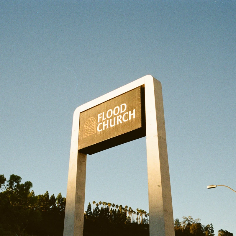 A low-angle, film-style photo of a large, modern sign for Flood Church against a clear blue sky, with trees and a street lamp in the background.