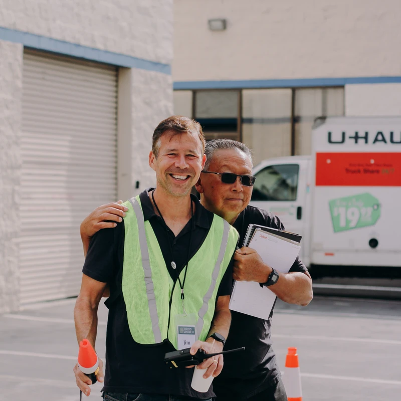 Two men smile for the camera in a parking lot. One wears a yellow safety vest, while the other has his arm around him and holds a clipboard.
