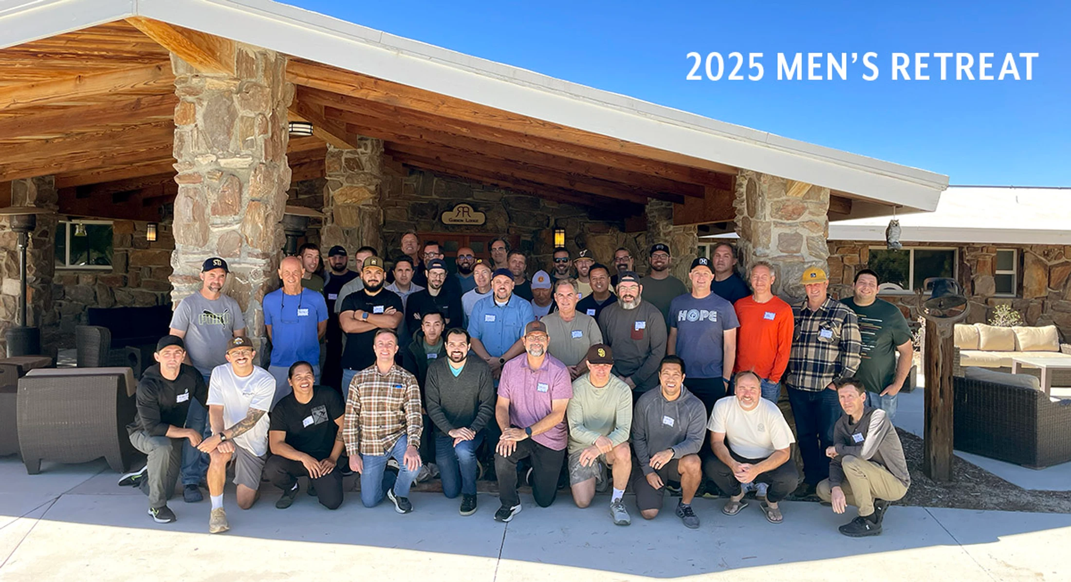 A large group of men posing for a photo outdoors in front of a rustic lodge under a clear blue sky, with text that reads '2025 MEN'S RETREAT'.