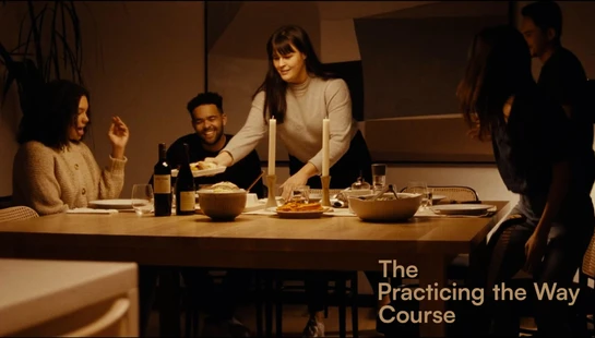 A diverse group of friends enjoys a candlelit dinner party. A woman serves food from a platter as others laugh and talk around a wooden table.