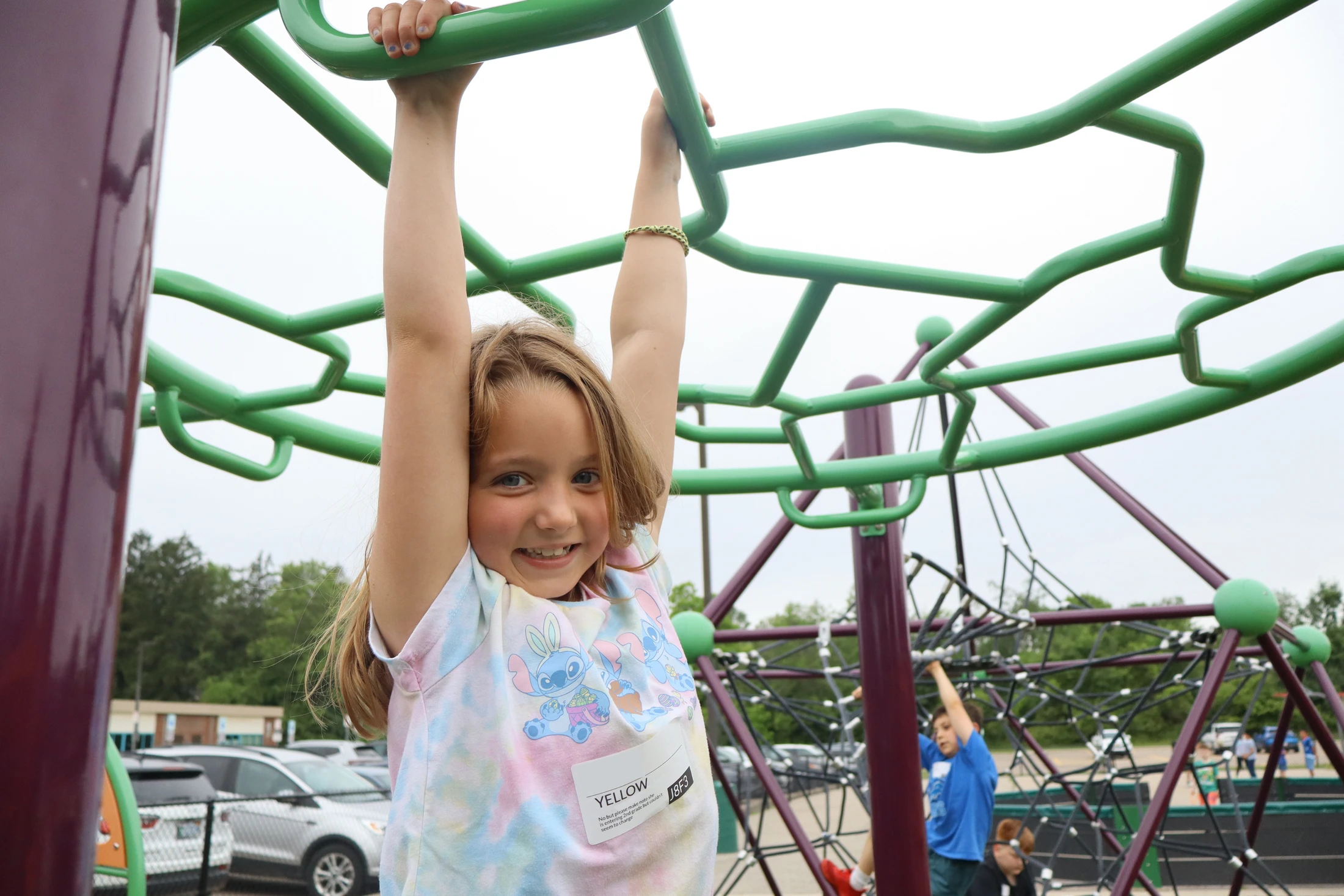 A young girl with blonde hair smiles at the camera while hanging from a green set of monkey bars on a school playground.