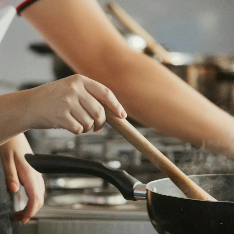 A close-up shot of a person's hands holding a wooden spoon, stirring food in a steaming black pan set on a gas stove with a blue flame.