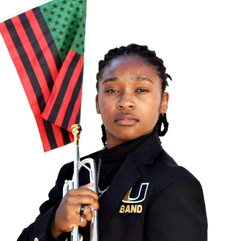 A young Black person in a band uniform looks seriously at the camera, holding a silver trumpet with a red, black, and green Pan-African flag attached.