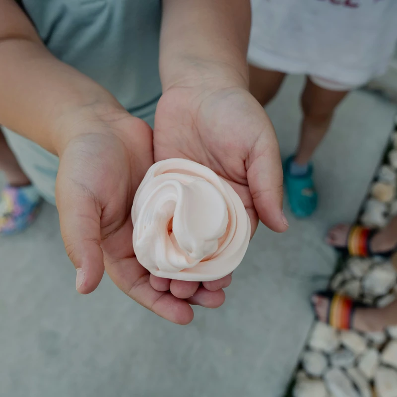 A top-down view of a child's cupped hands holding a swirl of fluffy, light pink slime. The feet of other children are out of focus in the background.