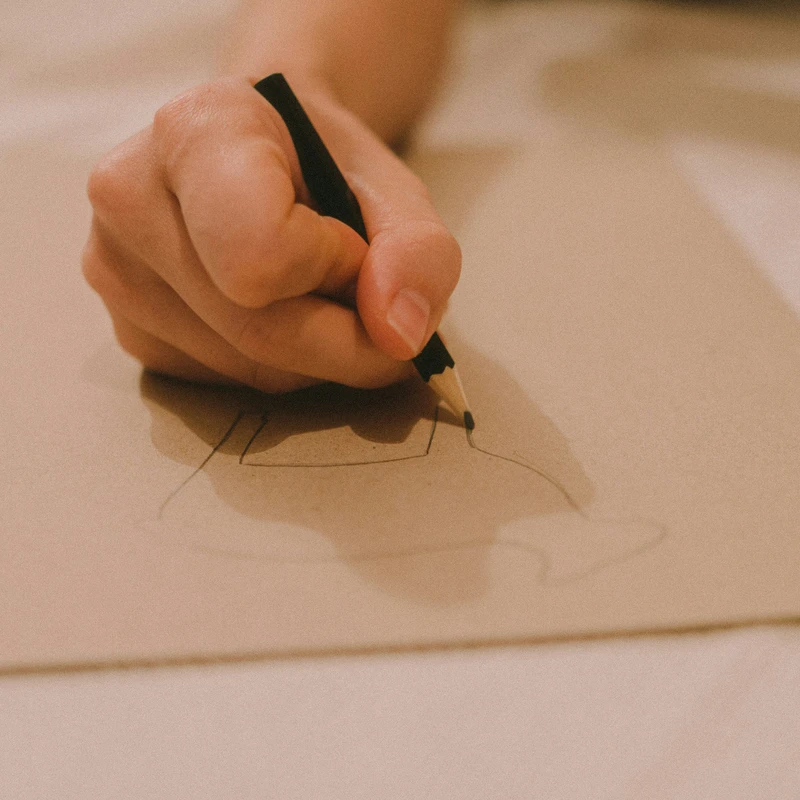 A close-up shot shows a person's hand holding a black pencil, sketching the outline of an object onto a sheet of light brown paper.