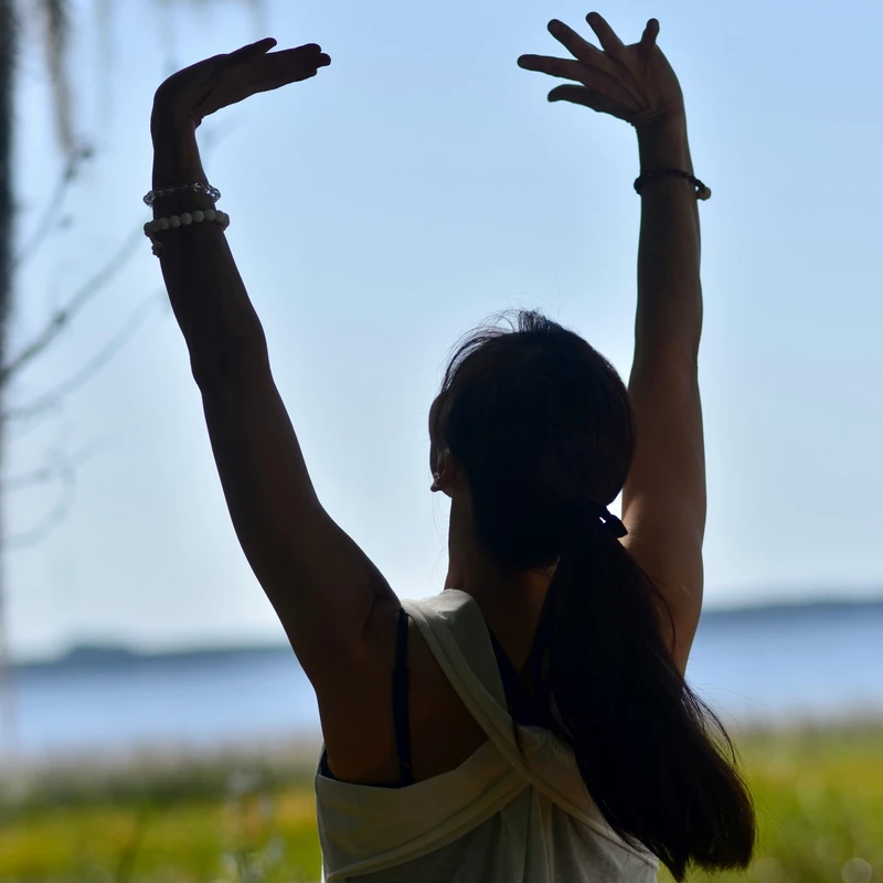 A woman seen from behind in silhouette, her arms raised towards the blue sky. She stands in a natural landscape with greenery and water in the background.