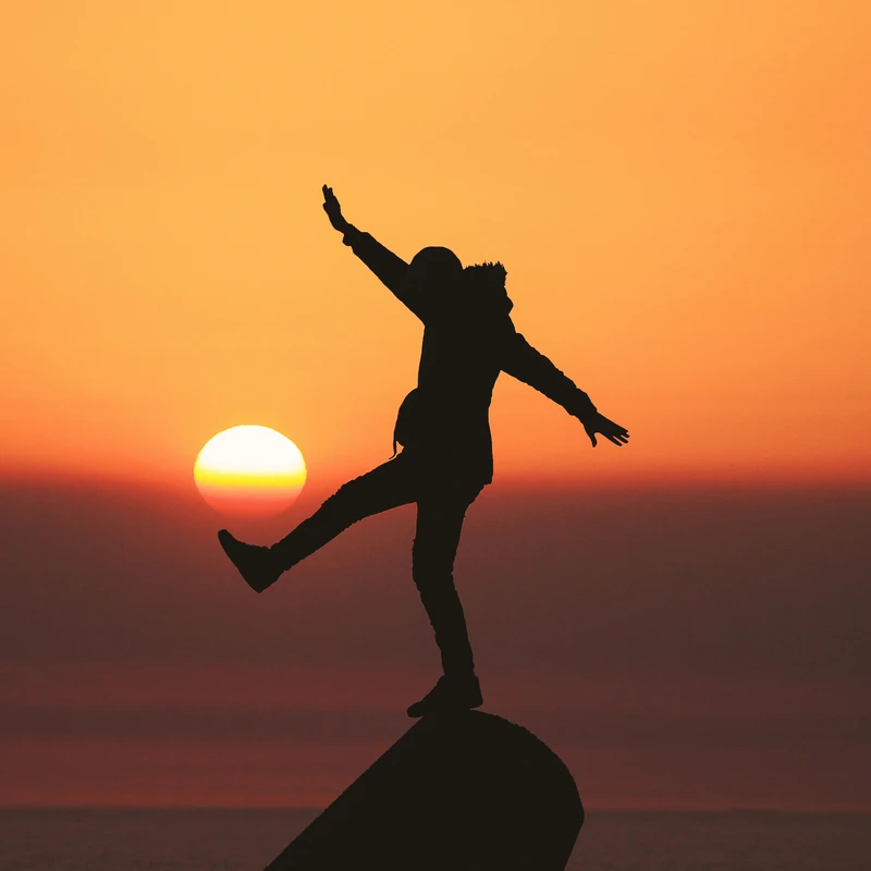 A silhouette of a person balancing on one leg against a vibrant orange sunset, with their other leg raised as if kicking the sun on the horizon.