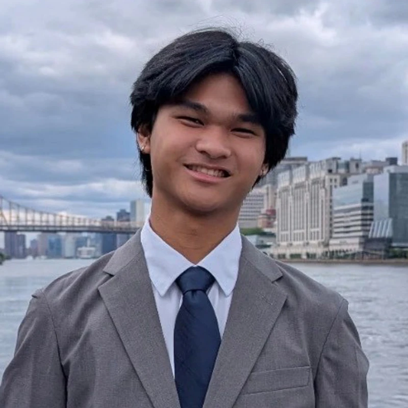 A young man with dark hair smiles while wearing a grey suit and tie. He is standing outdoors with a city skyline, a river, and a large bridge behind him.