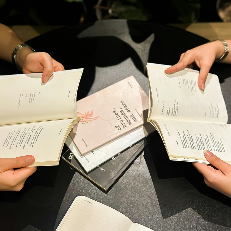 An overhead shot of two people sitting at a dark table, each holding open a book of poetry. More books are stacked between them.