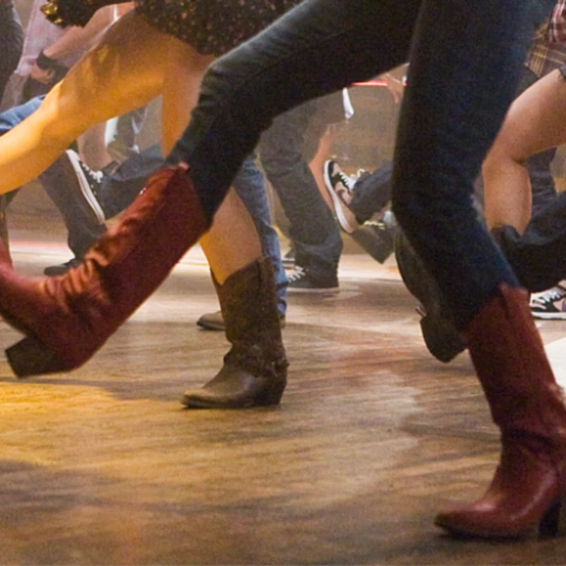 A low-angle shot of people line dancing. The focus is on their legs and feet, showing a variety of cowboy boots and jeans on a wooden dance floor.