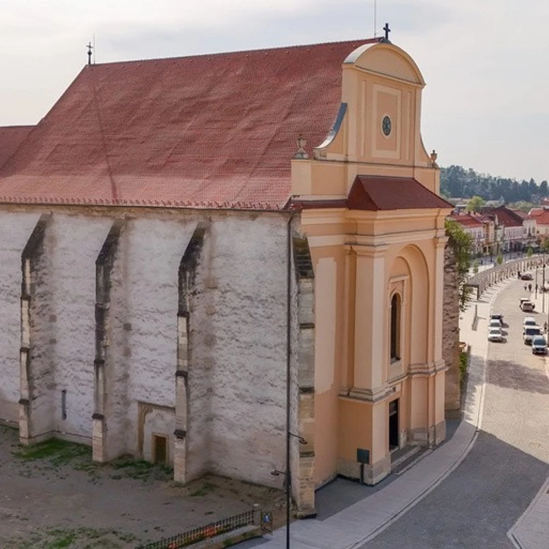 An elevated view of a large, historic church with a red tile roof and stone buttresses, next to a curving street in a European town.
