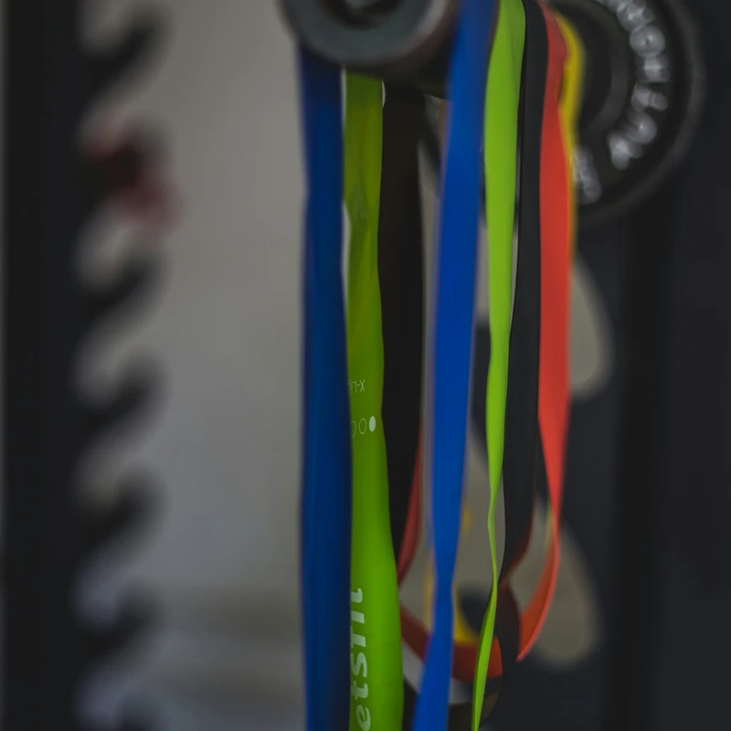 A close-up, selective focus shot of colorful resistance bands in blue, green, black, and red hanging from a metal bar in a gym.