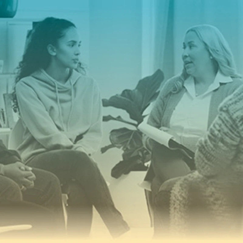 A diverse group of people sitting in a circle for a support group. A woman with a clipboard is speaking to the group, who are listening intently.