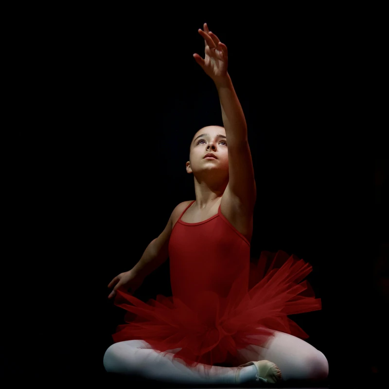 A young ballerina in a red leotard and tutu sits on a dark stage, lit from above. She looks up with a focused expression, reaching one arm towards the light.
