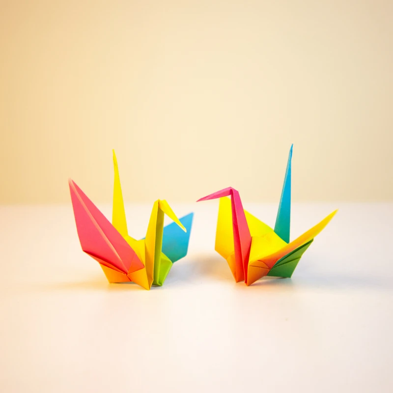 Two brightly colored origami cranes made from pink, yellow, blue, and green paper sit facing each other on a white surface against a plain beige background.