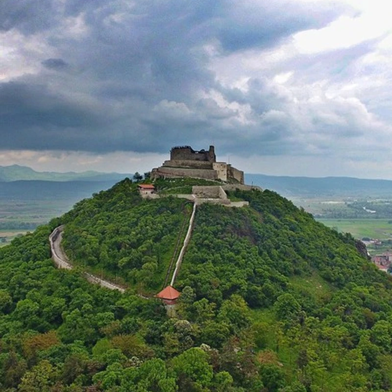 A stone fortress in ruins crowns a lush, green hill under a sky full of dark, dramatic clouds. A steep path and a winding road lead up the hillside.