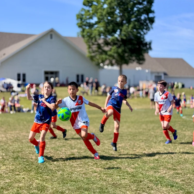A group of young children in soccer uniforms run on a grassy field, competing for a bright green ball during a game under a clear blue sky.