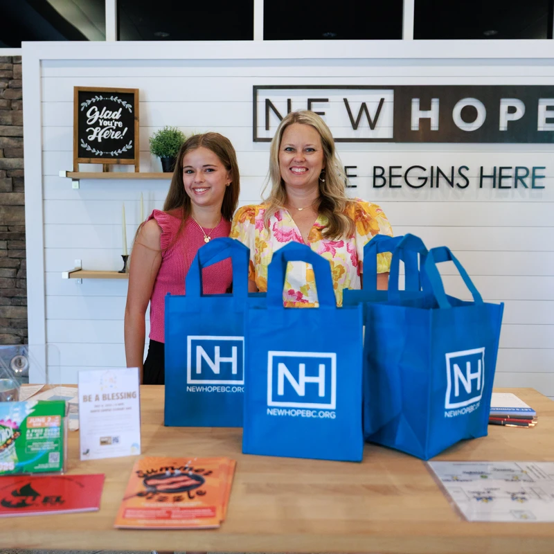 A smiling woman and a young girl stand behind the New Hope welcome desk, which has pamphlets and blue tote bags ready for visitors.