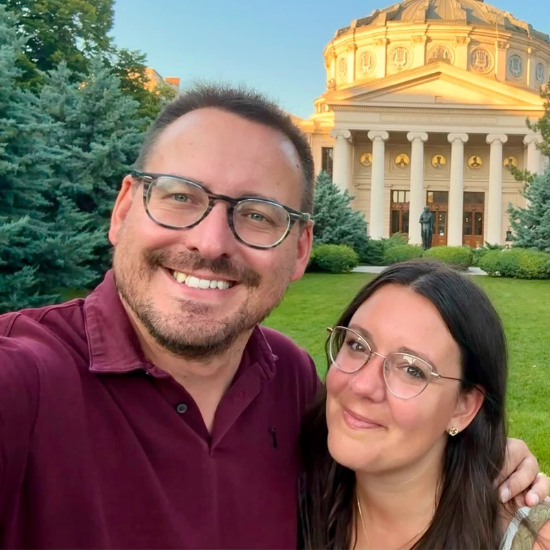A man and a woman, both wearing glasses and smiling, take a selfie on a green lawn in front of a grand, domed building with columns at sunset.