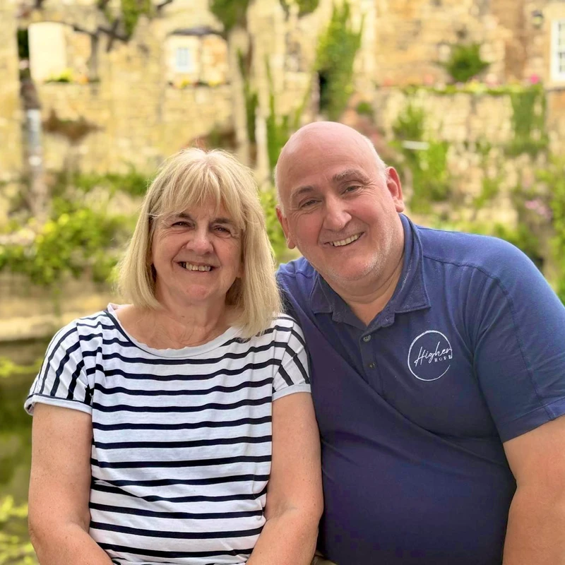 A smiling woman in a striped shirt and a man in a blue polo shirt sit together outdoors. In the background is a stone building next to a body of water.