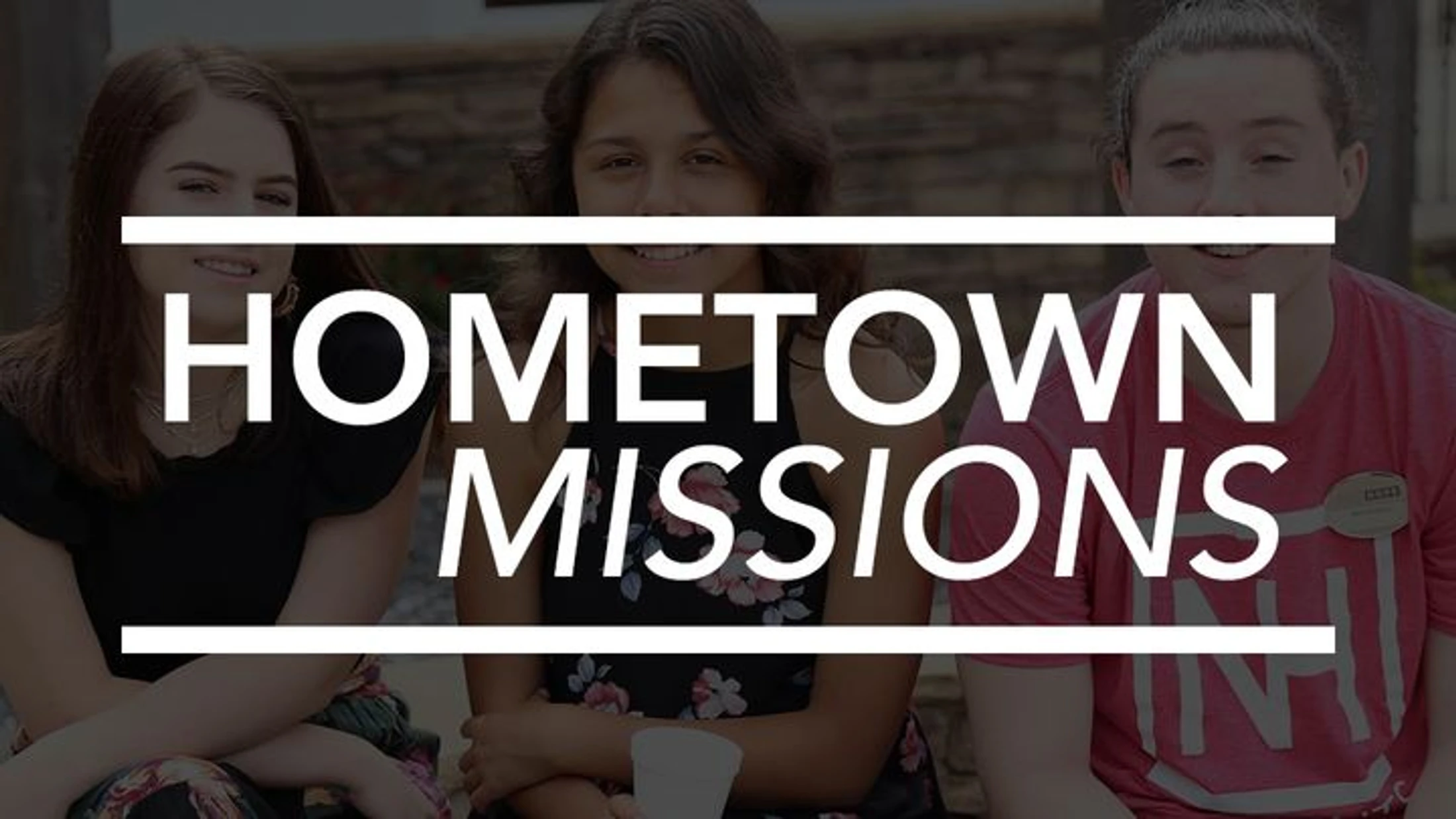 Three young women smile at the camera while sitting together, with the words 'Hometown Missions' in large white text overlaid on the image.