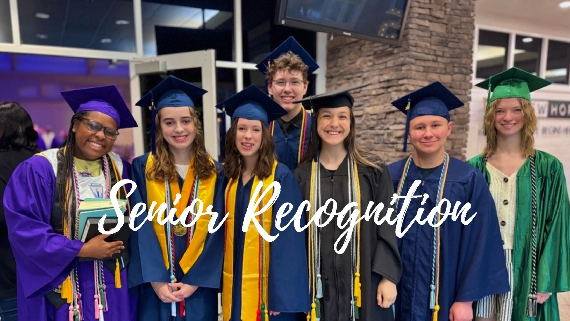 A group of seven diverse high school seniors in colorful caps and gowns smile for a group photo at their recognition ceremony.