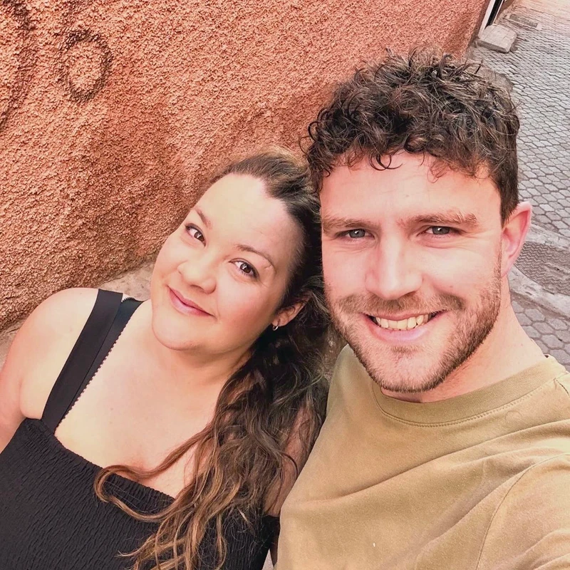 A smiling man and woman take a selfie together. The man has curly brown hair and the woman has long wavy hair. They are standing next to a textured orange wall.