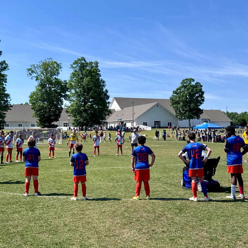 Two youth soccer teams stand on a grassy field under a clear blue sky. The team in the foreground wears blue jerseys and red shorts, facing the opposing team.